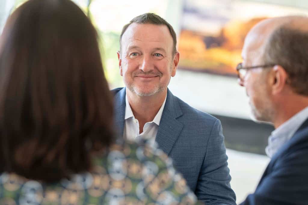 Man in a blue suit smiling during a conversation with two other people in a bright room.